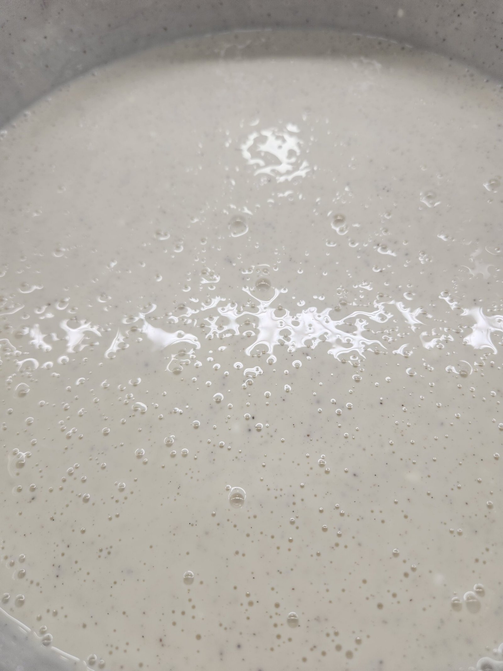 close-up da superfície do creme quente na panela, fluida e coberta de bolhas, escorrendo sem estrutura (substituto aproximado: brigadeiro de baunilha em cozimento — mesmo contraste de viscosidade quente vs. fria)