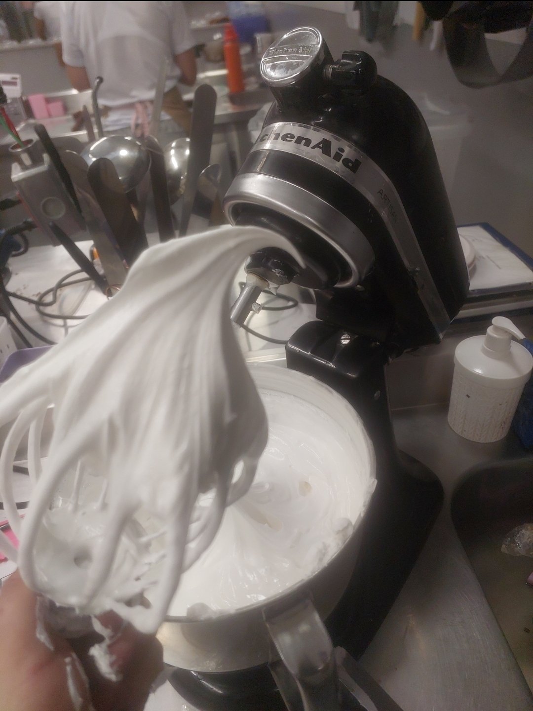 batedor retirado da tigela com massa branca brilhante formando pico volumoso, batedeira e tigela ao fundo (substituto aproximado: merengue italiano, não marshmallow — mesma calda quente vertida em espuma sob batimento)